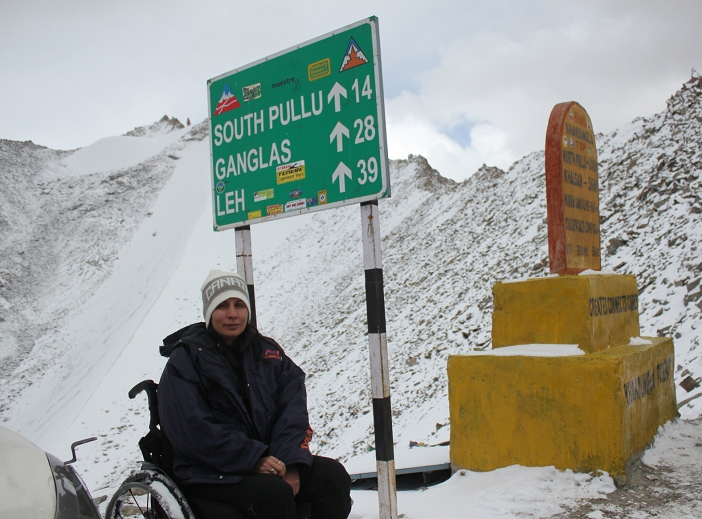 Sunita at Khardungla Pass on her wheelchair with snow mountains behind her and a direction board to Leh