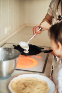 The hands of a mother and child pouring white batter from a ladle on to a pan indicative of making pancakes