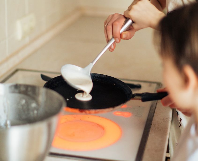 The hands of a mother and child pouring white batter from a ladle on to a pan indicative of making pancakes