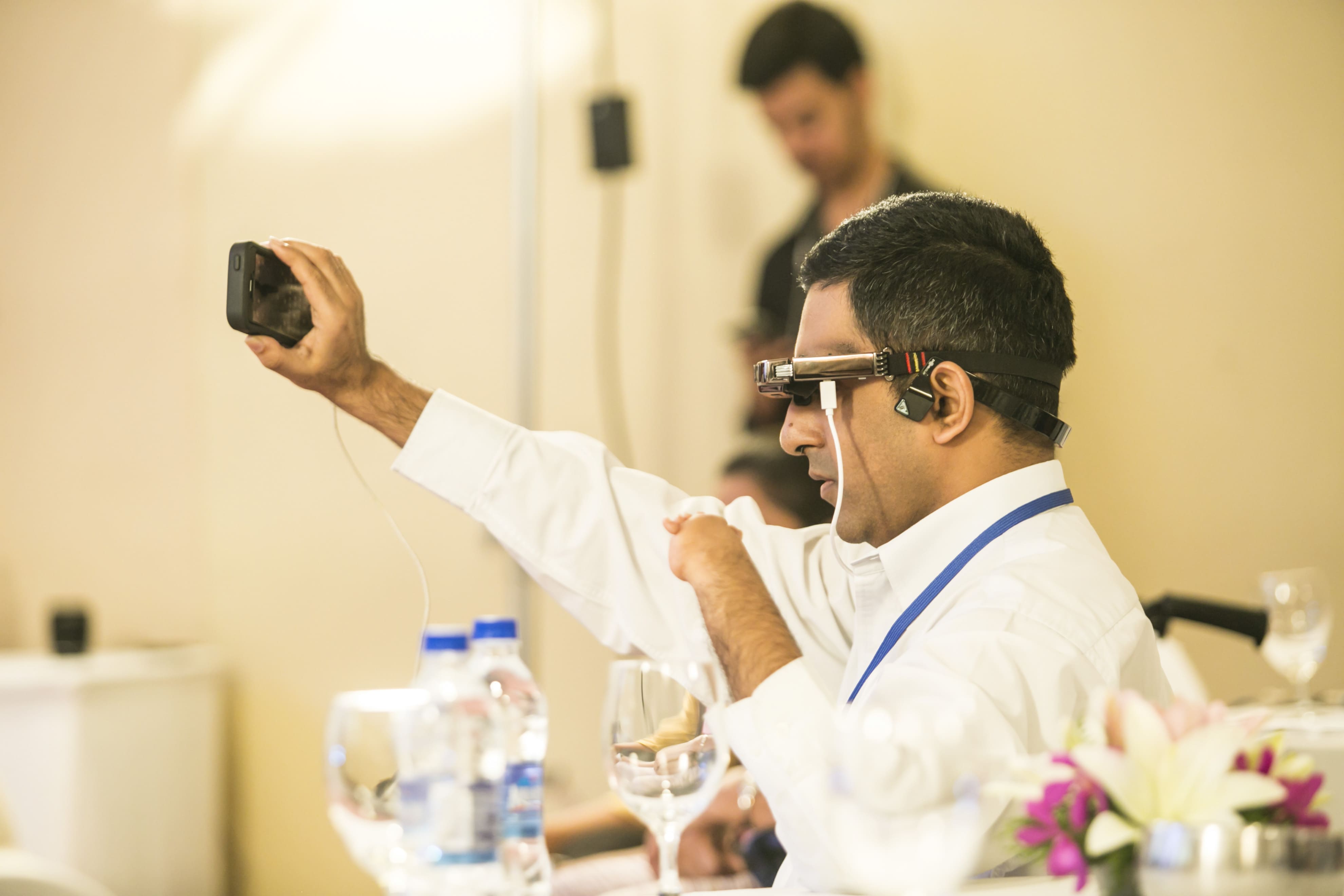 The image is the left-side view of a blind person holding a smartphone in his right hand in front of his head. He has an underdeveloped left arm and has an assistive device held on his ears connected to the phone.