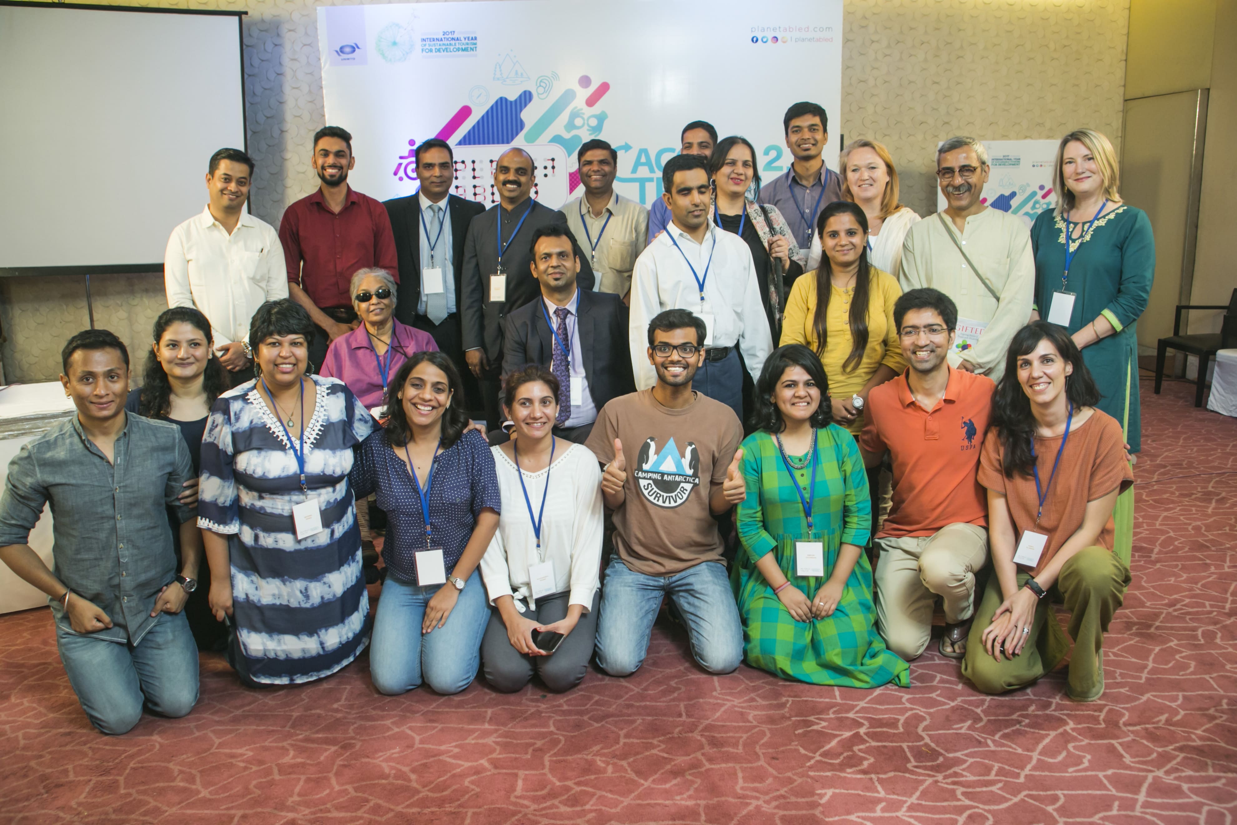 It is a group picture of about 25 people from diverse genders, age groups, racial backgrounds, ethnicities, and disabilities. They seem to have just participated in a summit or a formal meeting inside a convention hall.