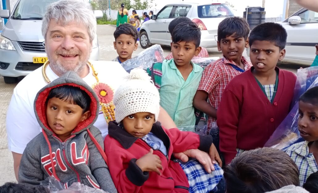 The image shows a group of children surrounding an elderly man in a wheelchair. The man is giving them a group hug and they all are smiling.