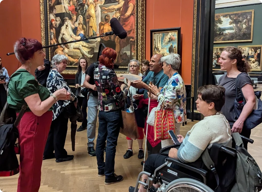 Inclusive museum tour with wheelchair users, deaf, blind and sighted visitors engaging in art discussion inside a gallery with classical paintings with a guide and sign language interpreter.
