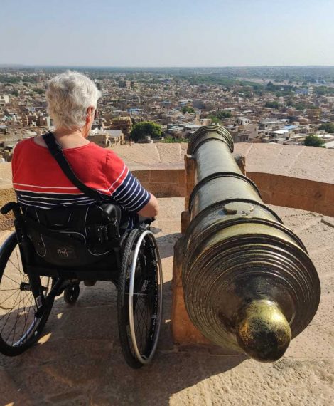 A wheelchair traveller is seen from the back. She is dressed in red and has short white hair. She is watching the city from the top of an old fort. A brass mental canon is placed directing towards the city.