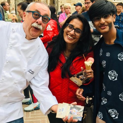 An old bald man with mustaches is seen smiling in a chef coat. He is goofing with an asian mother and teenage son duo with ice cream in their hands.